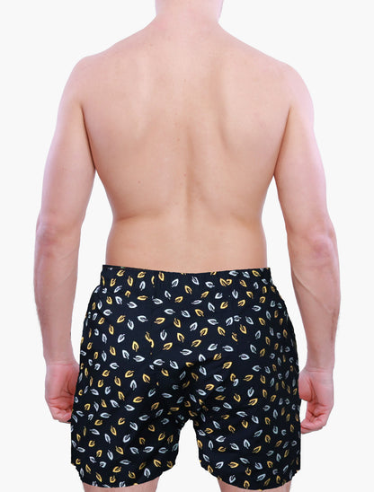 Back side of male model wearing black swim trunks with yellow and white leaf pattern on a plain background