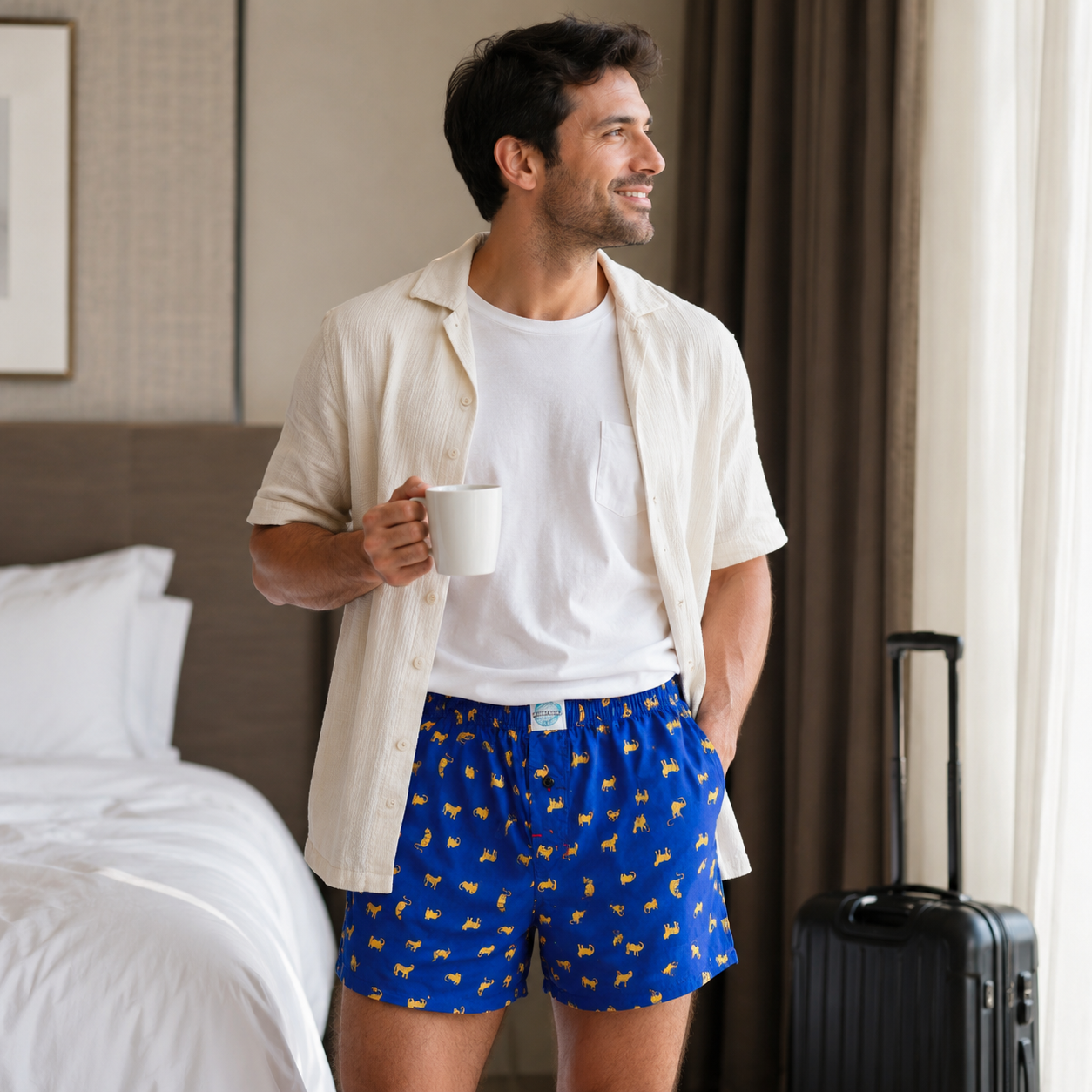 Man in a hotel room wearing blue boxer shorts with yellow patterns, holding a white mug.