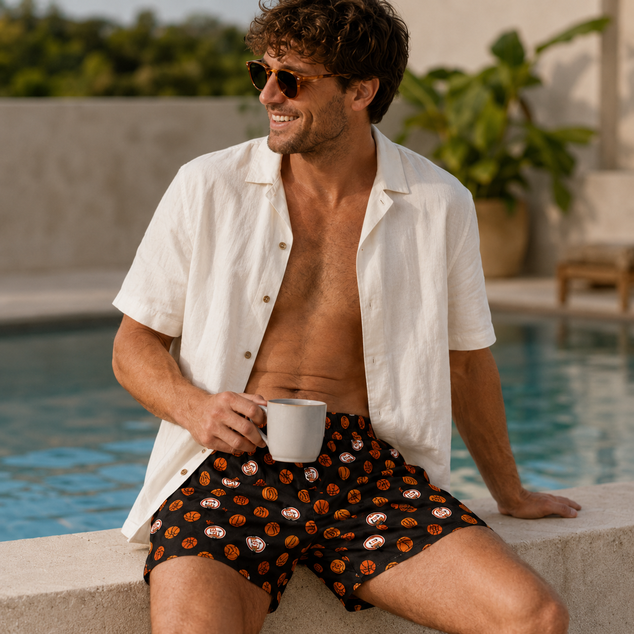Man in white shirt and Weunderlust boxer shorts sitting by a pool holding a cup.